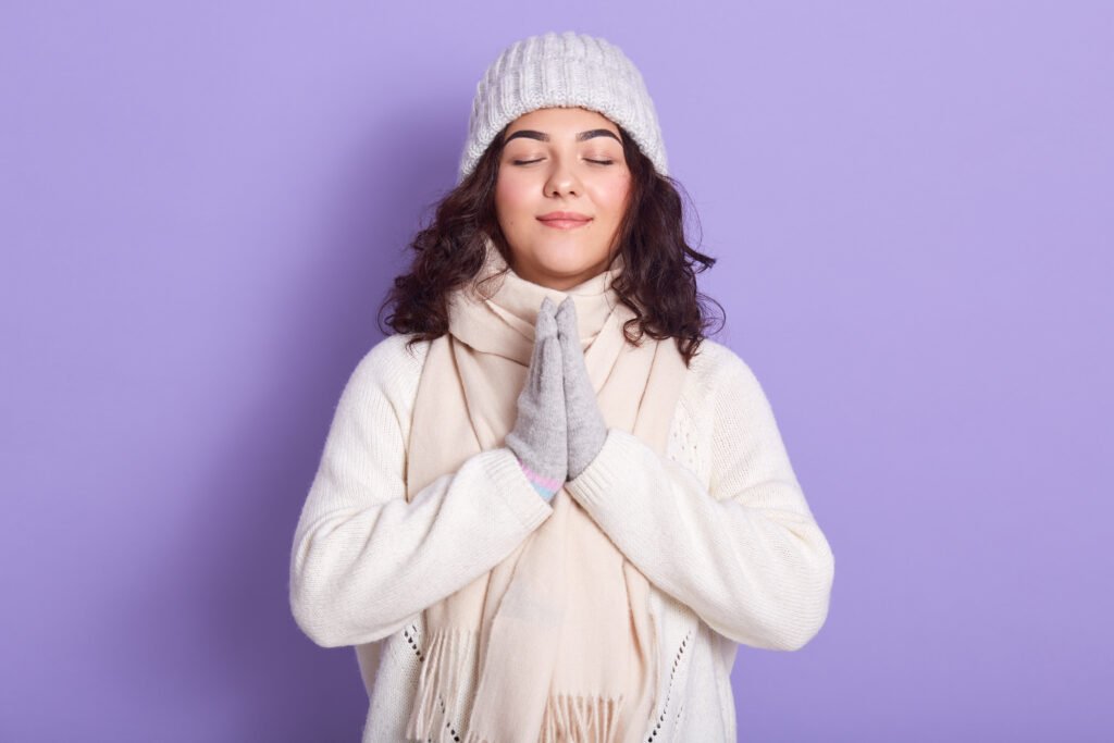 Winter Wellness Tips : Stay Active and Breathe Smart peaceful attractive young girl standing isolated over lilac background in studio, keeping hands together, closing eyes, having dark hair, wearing white sweater, scarf and hat. people and rest concept.
